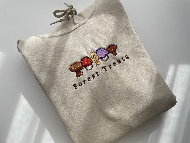 Może przedstawiać: Beżowa bluza z kapturem ze sznurkiem i haftowanym wzorem "Forest Treats". Wz&oacute;r przedstawia pikselowane grzyby w kolorze czerwonym, brązowym, fioletowym i ż&oacute;łtym. Bluza z kapturem jest złożona, pokazując prz&oacute;d z haftem.