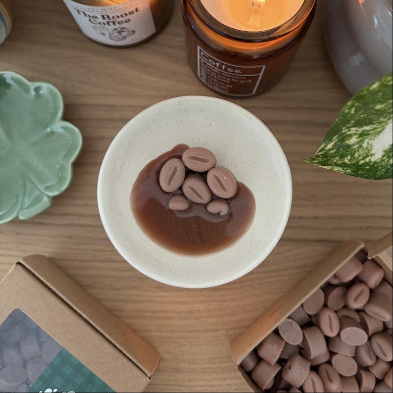 Op de afbeelding: Een klein, gebroken wit keramisch schaaltje met een bruine, koffie-achtige substantie en verschillende bruine, koffieboonvormige wassmelt. Twee kleine kartonnen dozen gevuld met vergelijkbare wassmelt zijn ook zichtbaar, samen met twee brandende kaarsen.