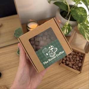 Op de afbeelding: Een vierkante, bruine kartonnen doos met een doorzichtig venster, gevuld met donkerbruine, vierkante lekkernijen. Een groen label met de tekst "The Roost Coffee" en koffie-thema graphics staat op de voorkant. Een andere doos met vergelijkbare lekkernijen is in de buurt open.