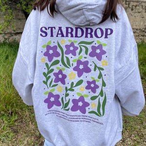 May include: A grey hoodie with a purple and green floral design and the text "STARDROP" in purple. The text below the design reads "A mysterious fruit that empowers those who eat it. The flavour is like a dream... a powerful personal experience, yet difficult to describe to others."