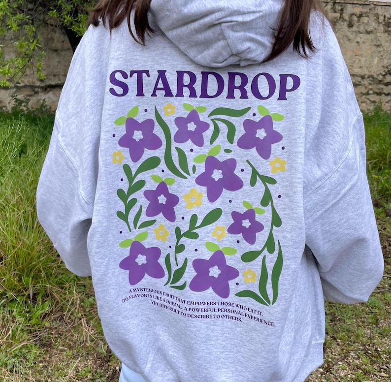 May include: A grey hoodie with a purple and green floral design and the text "STARDROP" in purple. The text below the design reads "A mysterious fruit that empowers those who eat it. The flavour is like a dream... a powerful personal experience, yet difficult to describe to others."