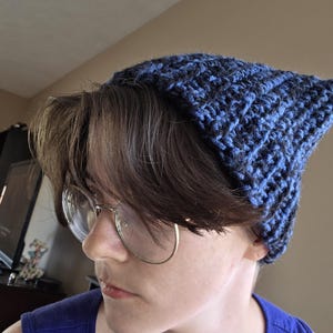 May include: A hand-crocheted beanie featuring a textured, ribbed pattern in a variegated blend of deep navy blue and black yarn. The hat has a slightly pointed, structured shape at the top and is crafted with a thick, warm, and durable yarn construction.