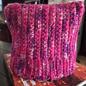 May include: A hand-crocheted beanie hat featuring a vibrant hot pink yarn base with subtle, variegated flecks of white, purple, and blue throughout. The hat is constructed with a vertical ribbed stitch pattern, creating a textured, cylindrical shape.