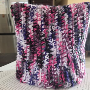 May include: A hand-crocheted beanie hat featuring a variegated yarn pattern in shades of bright pink, deep purple, black, and white. The accessory is constructed with a vertical ribbed stitch texture, creating a dense and warm fabric appearance.
