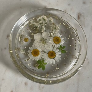 May include: A clear glass coaster with white flowers and gold glitter embedded in the resin. The flowers include daisies and baby's breath.