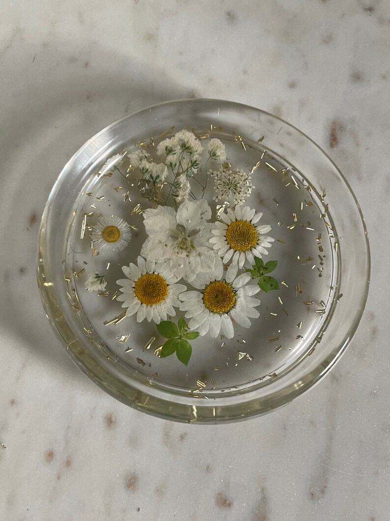 May include: A clear glass coaster with white flowers and gold glitter embedded in the resin. The flowers include daisies and baby's breath.