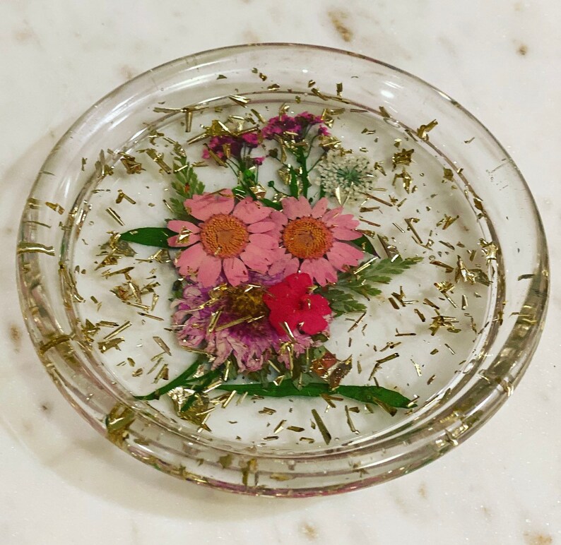 May include: A clear glass coaster with a floral design. The coaster features pink and red flowers, green leaves, and gold glitter.