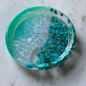 May include: A round, clear resin coaster with a light blue and white ombre design. The coaster is filled with small, turquoise-colored stones.
