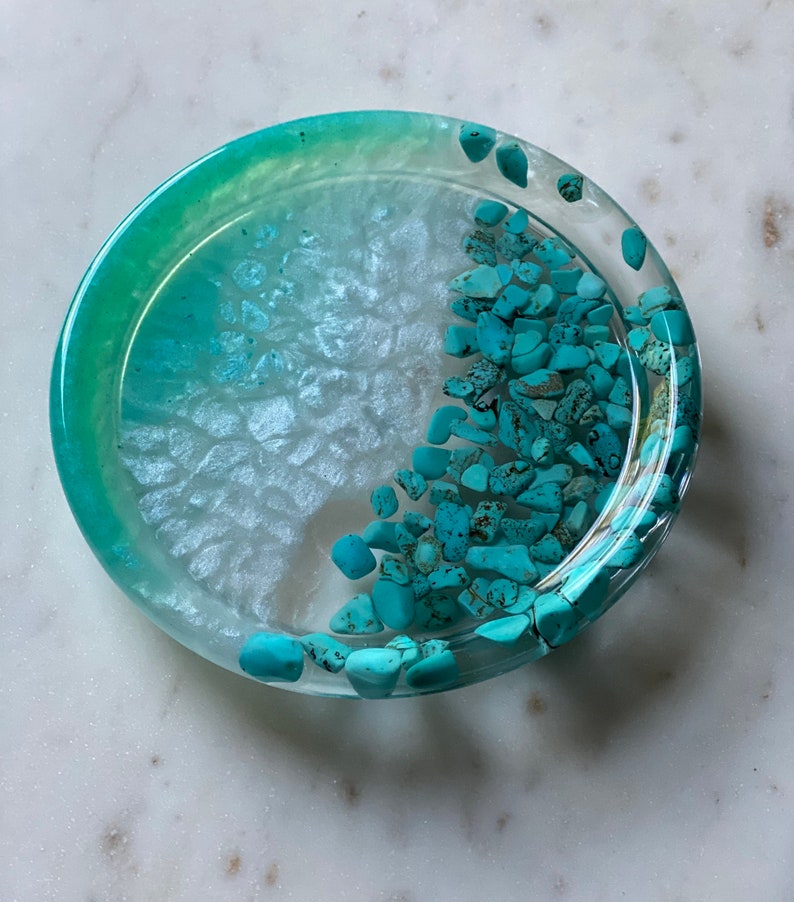 May include: A round, clear resin coaster with a light blue and white ombre design. The coaster is filled with small, turquoise-colored stones.
