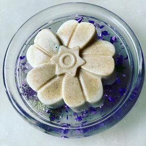May include: A white flower-shaped soap bar with a star design in the center, resting on a clear glass dish with purple glitter.