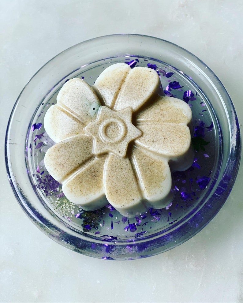 May include: A white flower-shaped soap bar with a star design in the center, resting on a clear glass dish with purple glitter.