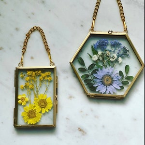May include: Two gold-toned picture frames with chains hanging from the top. The frame on the left is rectangular and contains pressed yellow flowers. The frame on the right is hexagonal and contains pressed purple and white flowers.