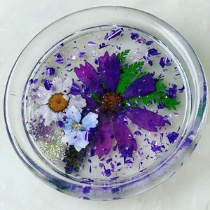 May include: A clear glass coaster with a purple, white, and blue pressed flower design. The coaster is filled with purple glitter.
