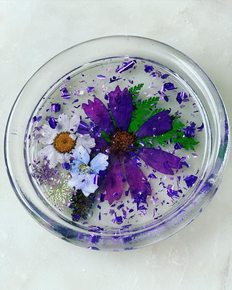 May include: A clear glass coaster with a purple, white, and blue pressed flower design. The coaster is filled with purple glitter.