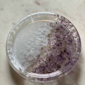 May include: A clear glass coaster with a white and purple design. The coaster is filled with small purple crystals.