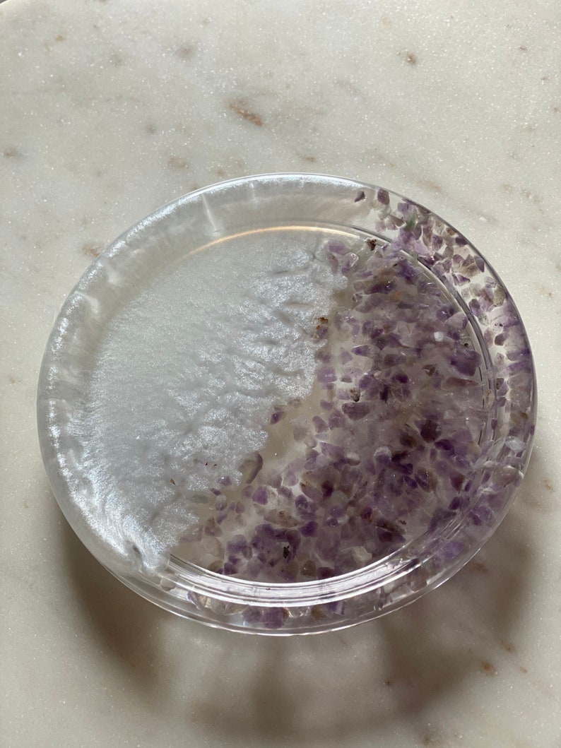 May include: A clear glass coaster with a white and purple design. The coaster is filled with small purple crystals.