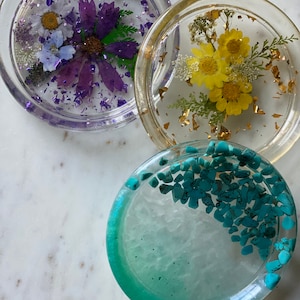 May include: Three round resin coasters with different designs. The first coaster features a purple flower with gold glitter. The second coaster features a yellow flower with gold leaf accents. The third coaster features a turquoise blue and white design with small turquoise stones.