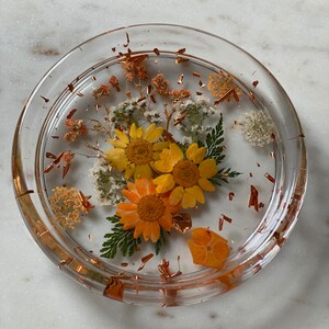 May include: A clear glass coaster with a layer of resin containing pressed flowers and copper leaf accents. The flowers are yellow, orange, and white.