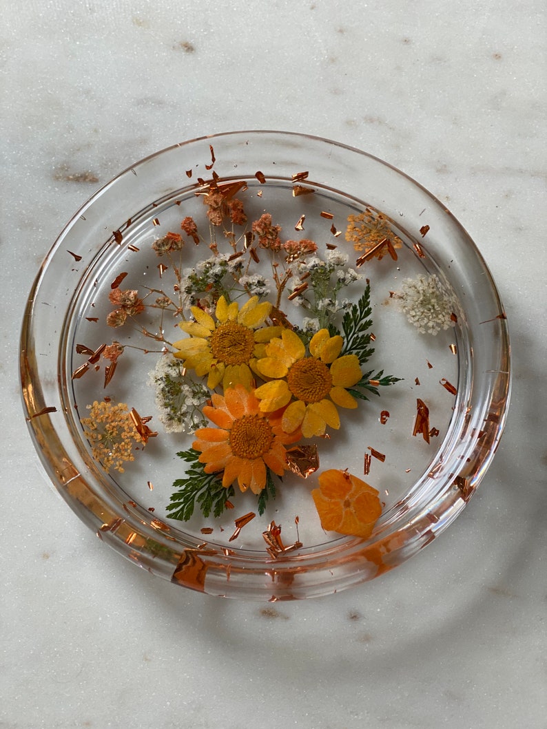 May include: A clear glass coaster with a layer of resin containing pressed flowers and copper leaf accents. The flowers are yellow, orange, and white.