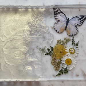 May include: A clear rectangular tray with a white butterfly and pressed flowers. The flowers are white and yellow and have green leaves.