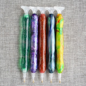 May include: Five diamond painting pens with different colored handles. The pens have clear plastic tips and are arranged in a row on a gray textured surface.