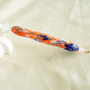 May include: A close-up of a colorful diamond painting pen with a white, orange, blue, and gold marbled design. The pen is lying on a beige fabric surface.