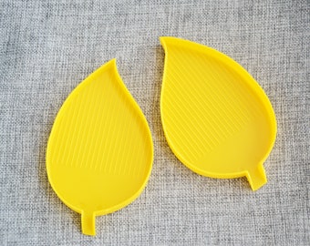 Puede incluir: Dos bandejas de color amarillo en forma de hoja con una superficie texturizada. Las bandejas son de plástico y están diseñadas para manualidades.