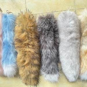 May include: A collection of six long, plush faux fur tail keychains in various colors including pastel pink, light blue, tan, dark gray, light gray, and golden brown, each featuring a silver-toned metal lobster clasp at the top end.