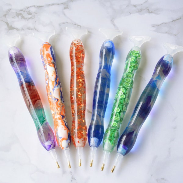May include: Six colorful diamond painting pens with clear handles and different colored resin tips. The pens have a variety of patterns and designs, including swirls, glitter, and lime slices.