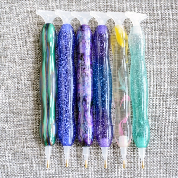 May include: Six diamond painting glue pens with different colored glitter and designs. The pens have clear plastic tips and are arranged in a row.