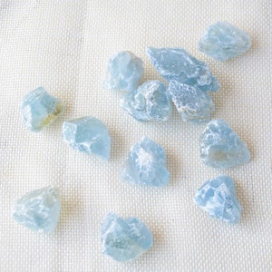 Può includere: Una collezione di pietre preziose di acquamarina azzurra, tagliate in modo grezzo. Le pietre variano per dimensioni e forma, con un aspetto leggermente traslucido. Sono sparse su un tessuto color crema, creando un'estetica naturale.