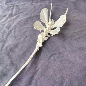 May include: A white, decorative wand with a floral design. The wand features a long, slender handle and a detailed, flower-like top with multiple petal-shaped elements. The object is set against a textured, gray-blue fabric.