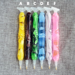 May include: Six diamond painting pens with different colored handles. The pens are labeled A through F. The pens are on a gray fabric background.