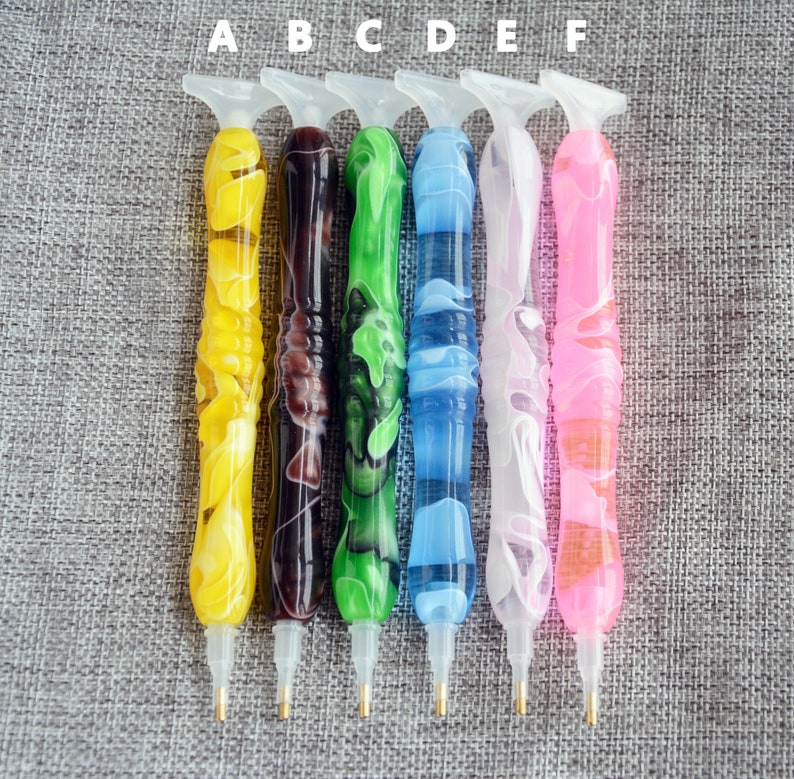 May include: Six diamond painting pens with different colored handles. The pens are labeled A through F. The pens are on a gray fabric background.