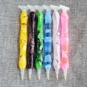 May include: Six diamond painting pens with different colored swirl designs. The pens have a clear plastic handle with a metal tip.
