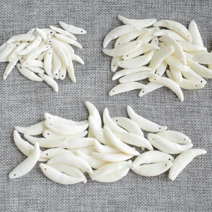 May include: A pile of white, crescent-shaped, polished seashells with a hole drilled through each one. The shells are arranged in three groups on a gray fabric background.