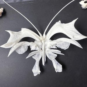 Op de afbeelding: Een delicate, witte vlindersculptuur met ingewikkelde vleugels. De vlinder staat tegen een donkere achtergrond, waardoor het gedetailleerde, getextureerde ontwerp opvalt. De antennes van de sculptuur steken omhoog.
