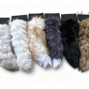 Puede incluir: Una colección de seis colas de piel sintética en varios colores, incluyendo gris, blanco, bronceado con punta blanca, gris oscuro con punta blanca, negro sólido y marrón con puntas negras. Cada cola está unida a una lengüeta rectangular negra.