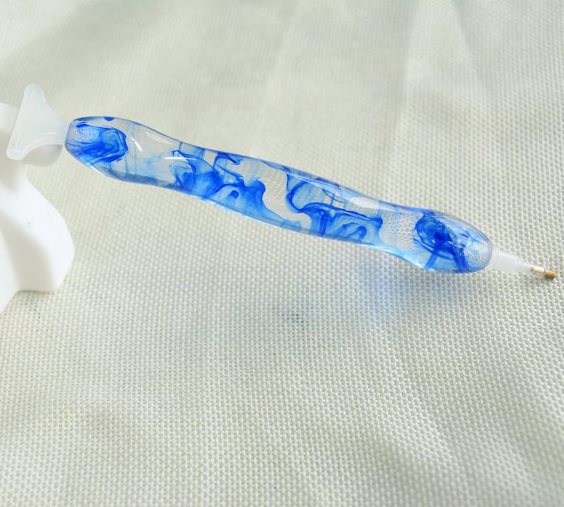 May include: A clear blue and white diamond painting pen with a swirl design. The pen has a gold tip and a white plastic handle.