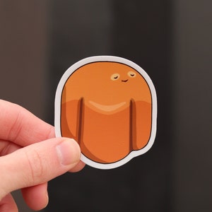 May include: A die-cut sticker featuring a smiling, cartoonish character in shades of orange and brown. The character has a rounded body with two small appendages and is outlined in white. The sticker is held by a hand against a dark background.