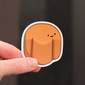 May include: A die-cut sticker depicting a cartoon character with a rounded orange body, two short legs, and a small smiling face. The sticker has a white border and is held by a hand against a dark background.