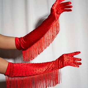 Peut inclure: Une paire de longs gants en satin rouge avec des franges et des strass rouges. Les gants sont montr&eacute;s sur les bras d'une personne.