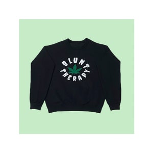 Op de afbeelding: Zwart sweatshirt met een witte en groene afbeelding van een marihuanablad en de tekst "Blunt Therapy" in een cirkel.