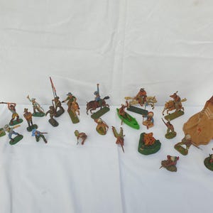 May include: A collection of vintage plastic toy cowboys and Native Americans, including a teepee, a canoe, and a small wooden building. The figures are painted in various colours, including brown, red, blue, and green.