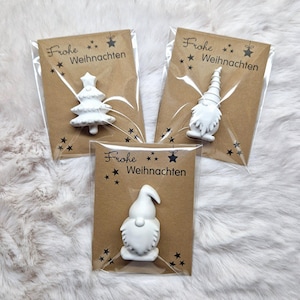 May include: Three small, white Christmas ornaments in clear plastic packaging, each attached to a brown card. The cards feature the text "Frohe Weihnachten" with star and snowflake designs. One ornament is a Christmas tree, and the other two are gnomes.