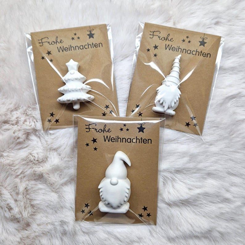 May include: Three small, white Christmas ornaments in clear plastic packaging, each attached to a brown card. The cards feature the text "Frohe Weihnachten" with star and snowflake designs. One ornament is a Christmas tree, and the other two are gnomes.