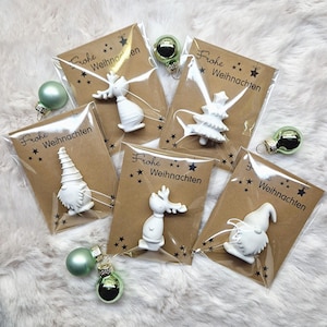 Könnte beinhalten: Eine Sammlung weißer Weihnachtsschmuckstücke in verschiedenen Formen, darunter ein Rentier, ein Wichtel und ein Weihnachtsbaum, verpackt auf braunen Karten mit dem Text "Frohe Weihnachten". Grüne Glaskugeln liegen um die Päckchen herum.