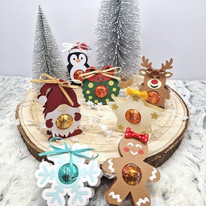 Könnte beinhalten: Eine Sammlung von sechs dekorativen Weihnachtsschmuckstücken aus Papier, die jeweils einen kreisförmigen Ausschnitt mit einer runden Lindt-Schokoladentrüffel enthalten. Die Motive umfassen Wichtel, Pinguin, Kranz, Rentier, Stern, Schneeflocke und Lebkuchenmann.