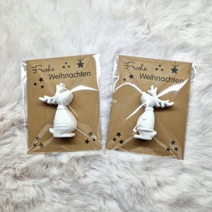 May include: Two white ceramic reindeer ornaments, each packaged on a brown card with the German text "Frohe Weihnachten" and star accents. The reindeer have detailed antlers and a festive design.
