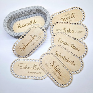 Könnte beinhalten: Ein grauer, ovaler Häkelkorb mit einem Holzsockel mit der Aufschrift "Kosmetik". Mehrere ovale Holzstücke mit dekorativem Text, darunter "Home Sweet Home" und "Carpe Diem". Die Holzstücke haben kleine Löcher an den Rändern.